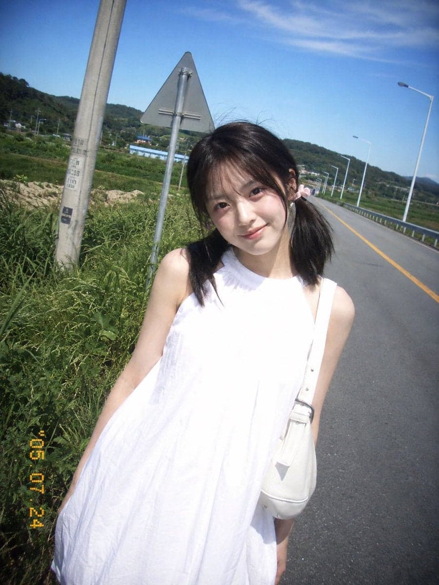 复古 CCD 卡片机夏日旅行抓拍,一位20岁出头的年轻东亚女idol站在乡间公路边。黑棕色低位双马尾,细碎刘海,浅粉色发